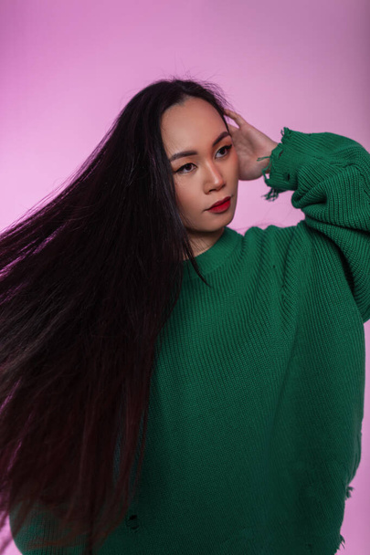 Fashion beautiful young Vietnamese model girl with black long hair in a fashionable green knitted sweater in the studio on a pink background - Фото, изображение