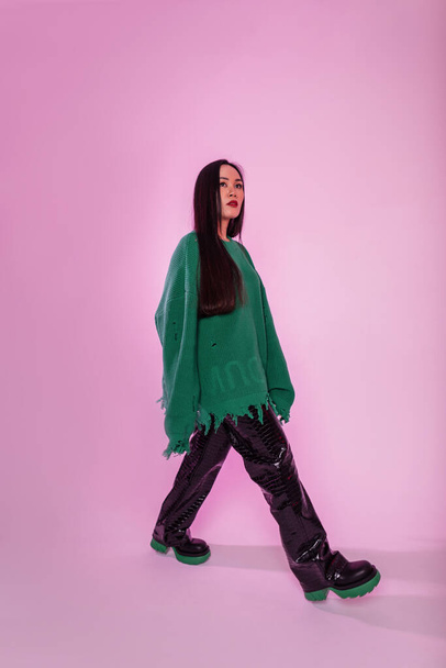 Μοντέρνο όμορφο κορίτσι σε ένα μοντέρνο πράσινο σκισμένο πουλόβερ με δερμάτινα παντελόνια και μπότες βόλτες στο στούντιο με ροζ φόντο - Φωτογραφία, εικόνα