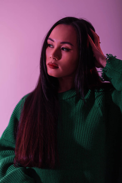 Stylish Asian model girl in fashionable green knitted sweater in studio with pink light. Color female portrait - 写真・画像