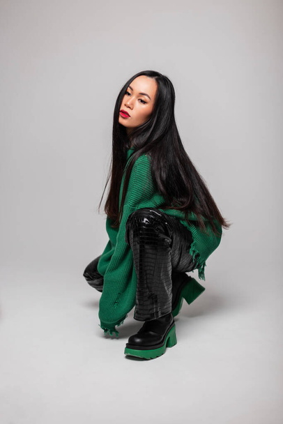 Fashionable beautiful young woman model with long hair in a fashionable green sweater with leather pants and leather boots sits and poses on a gray background - Foto, imagen