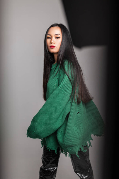 Μόδα Κορεάτισσα όμορφη γυναίκα με μακριά μαλλιά σε ripper κομψό πλεκτό πράσινο πουλόβερ και δερμάτινο παντελόνι βόλτες και ποζάρει στο στούντιο - Φωτογραφία, εικόνα