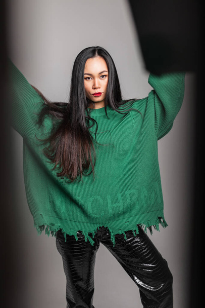 Stylish beautiful Chinese girl in fashionable clothes with ripped green sweater and leather pants poses in the studio - Photo, Image