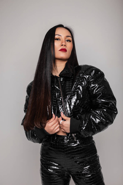Modieus mooi jong Aziatisch meisje met make-up en lang haar in een modieuze zwarte slangenhuid outfit met een jas en broek poseert in de studio tegen een grijze achtergrond - Foto, afbeelding