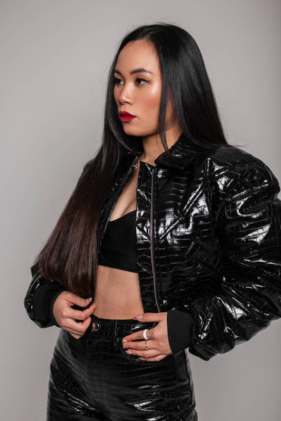 Fashionable beautiful sexy Asian woman in fashionable black snakeskin outfit with black jacket, bra and pants in the studio on a gray background - Zdjęcie, obraz