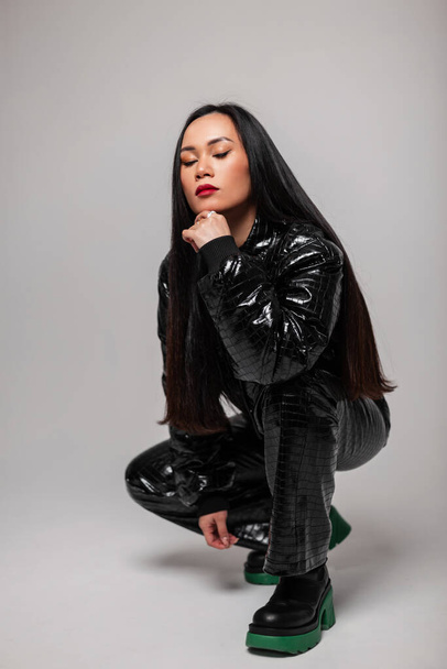Fashionable adorable Asian woman model in stylish snakeskin outfit with black stylish jacket, pants and boots poses in the studio - Photo, Image