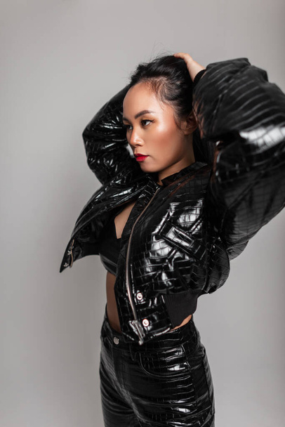 Cool glamourosa jovem mulher asiática em roupas da moda com jaqueta preta de couro, sutiã e calças posa no estúdio contra um fundo cinza - Foto, Imagem