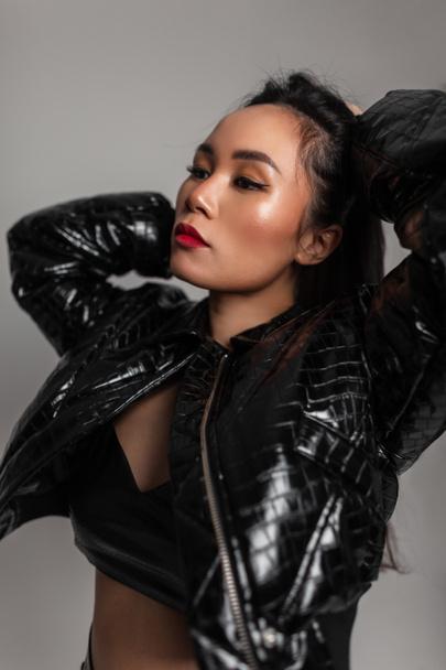 Elegante retrato asiático glamoroso de una hermosa joven de moda con maquillaje en una chaqueta de cuero negro y posando en el estudio sobre un fondo gris - Foto, Imagen