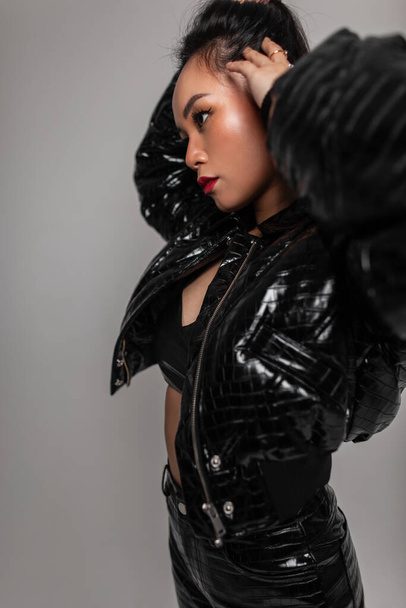 Fashionable beautiful Chinese girl in stylish leather with snakeskin jacket, black top and pants poses in the studio - Photo, image
