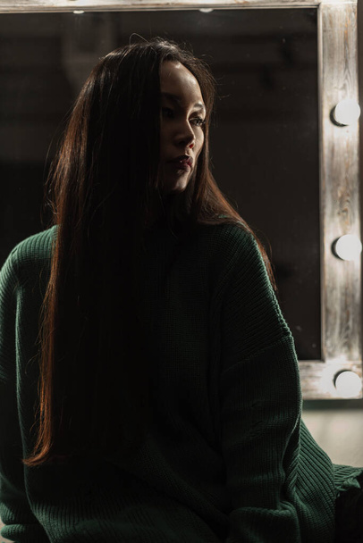 Beautiful Asian girl in a vintage green sweater sits in the dark near the mirror with light bulbs - Foto, imagen