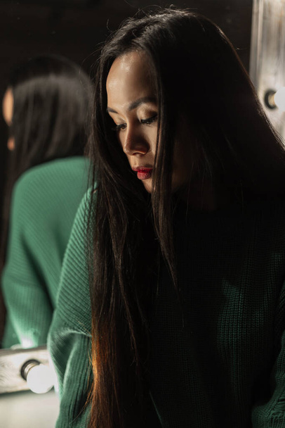 Stylish beautiful young girl with long hair in fashionable clothes with a vintage green knitted sweater in a dark room - Foto, imagen