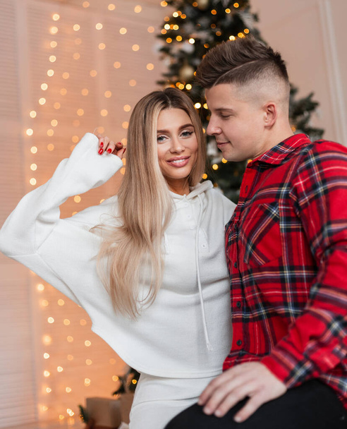 Happy young beautiful couple in fashionable clothes hugging and love at home near the lights. Fashion blonde woman and handsome guy indoors - Zdjęcie, obraz