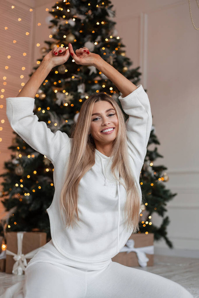 Feliz bela jovem loira com sorriso em roupas brancas senta-se e uma poses no chão perto de uma luz de Natal - Foto, Imagem