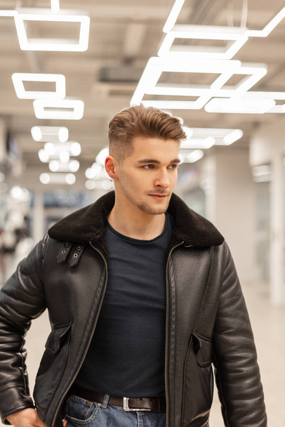 Fashionable hipster male model with hair in stylish clothes with a T-shirt and a black leather jacket with fur walks indoors with modern light - Фото, изображение