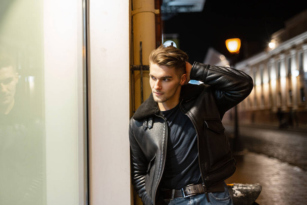 Modelo joven guapo de moda con peinado en una elegante chaqueta de cuero negro caminando en una ciudad nocturna y de pie cerca de un escaparate - Foto, Imagen