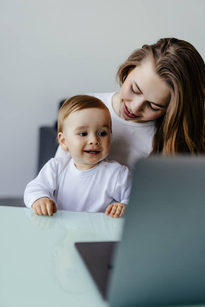 Jeune mère et son fils assis à table et utilisant un ordinateur portable à la maison. Maman souriante travaillant à la maison avec son enfant sur les genoux tout en écrivant un e-mail. - Photo, image