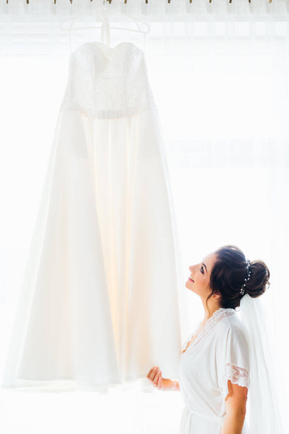 Panna młoda z piękną fryzurą patrzy na suknię ślubną na tle okna - Zdjęcie, obraz