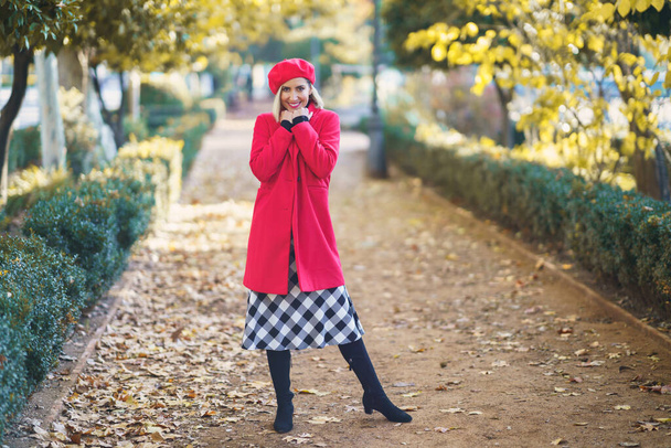 Teljes test vidám nő piros kabátban és svájcisapkában nézi a kamerát, miközben áll az ösvényen a parkban ősszel - Fotó, kép