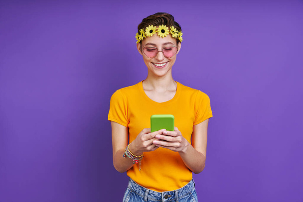Happy woman with floral head wreath using smart phone while standing against purple background - Φωτογραφία, εικόνα