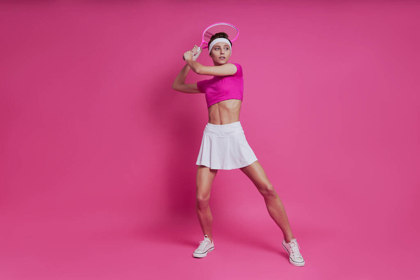 Confident young woman holding tennis racket while standing against pink background - 写真・画像