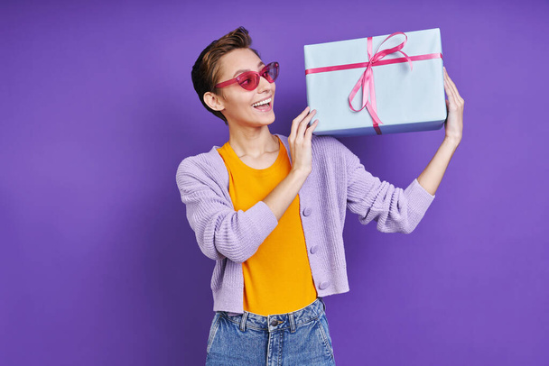 Happy young woman carrying gift box on shoulder while standing against purple background - Foto, Imagem