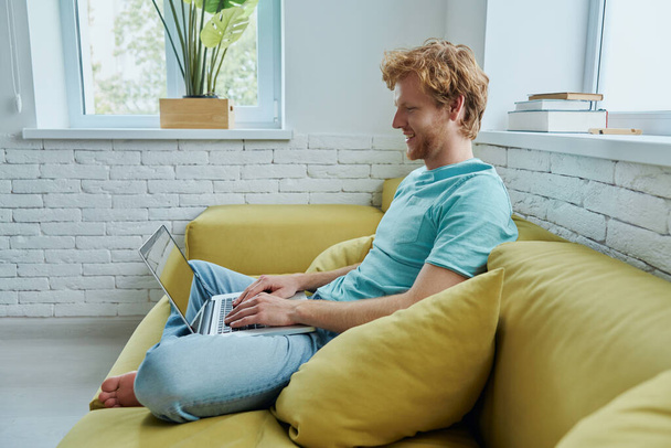 Confident redhead man using laptop and smiling while sitting on the couch at home - Fotografie, Obrázek