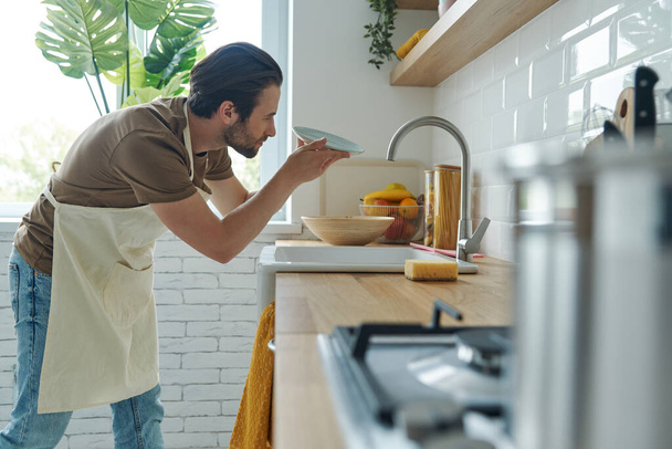 Confident young man checking dishes after washing while standing near kitchen sink - Foto, imagen