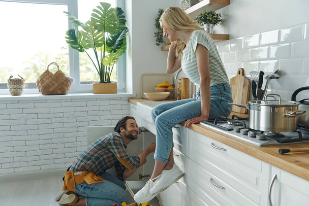 Cheerful young man repairing a sink while woman sitting on the kitchen counter - Φωτογραφία, εικόνα