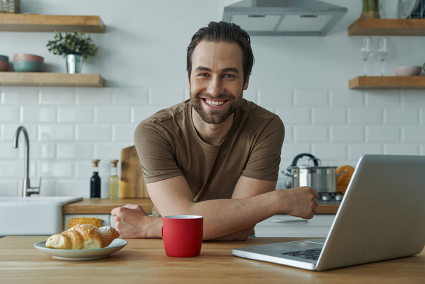 Fröhlicher Mann genießt das Frühstück und arbeitet am Laptop, während er auf der Kücheninsel sitzt - Foto, Bild