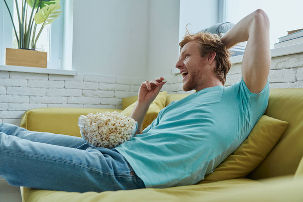 Šťastný mladý zrzka muž těší popcorn zatímco sedí na gauči doma - Fotografie, Obrázek