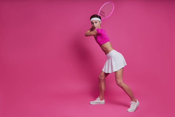Confident young woman holding tennis racket while standing against pink background - Fotografie, Obrázek