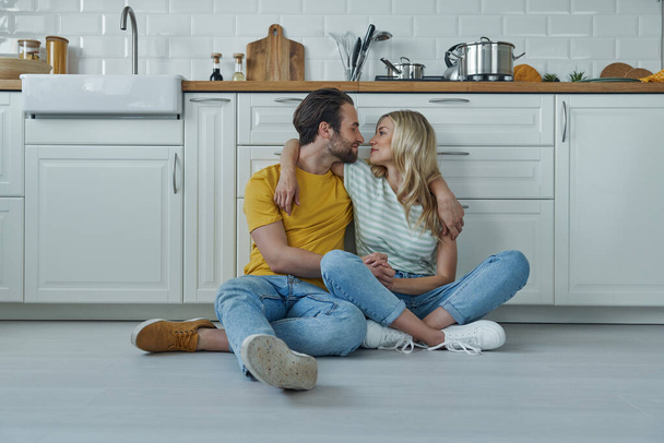 Beautiful young couple embracing while sitting on the floor and leaning at kitchen counter - Foto, imagen