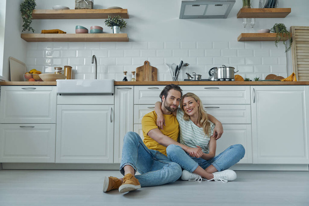 Beautiful young couple embracing while sitting on the floor and leaning at kitchen counter - 写真・画像