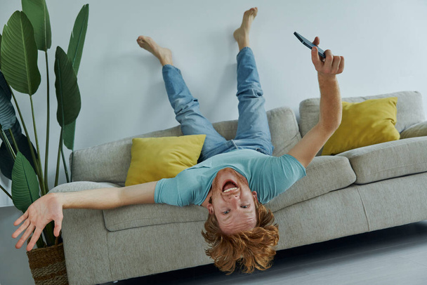 Playful redhead man holding smart phone and gesturing while relaxing on the couch at home - Фото, изображение