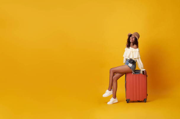 Młoda, ładna afrykańska turystka uśmiechnięta siedząca na bagażu z kamerą w ręku, odizolowana na żółtym tle. - Zdjęcie, obraz