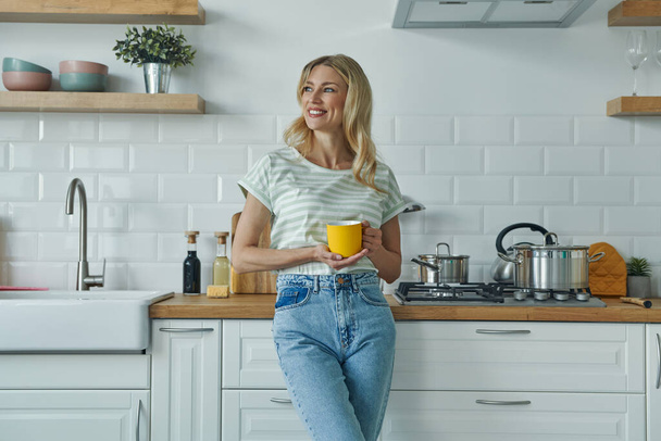 Cheerful young woman holding coffee cup and leaning at the kitchen desk - Photo, image