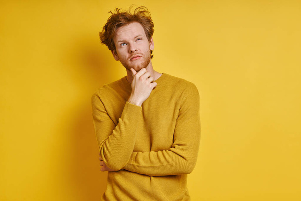 Thoughtful redhead man holding hand on chin while standing against yellow background - Foto, immagini