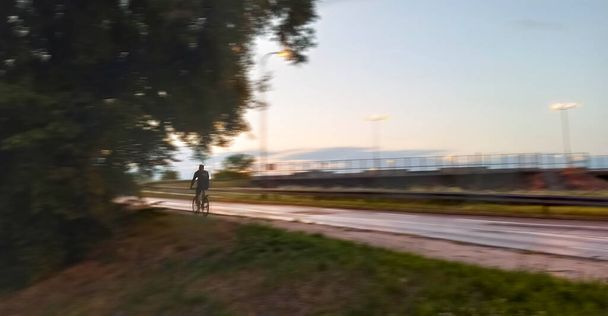 Motion blurred panning shot of a man riding a bicycle or bike on a side walk next to road and green field. healthy lifestyle concept - Photo, Image