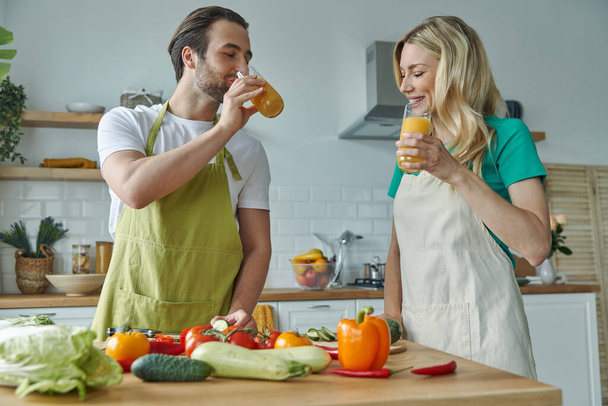 Happy couple drinking orange juice and smiling while cooking at the kitchen together - Fotografie, Obrázek