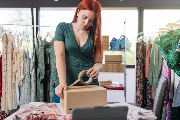 young business woman, closing a cardboard box with adhesive tape, order for customer of her online clothing shop. Young woman preparing a package for a client in the office. work and business concept - 写真・画像