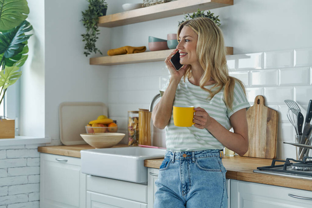 Attractive young woman talking on mobile phone while enjoying coffee at the kitchen - 写真・画像