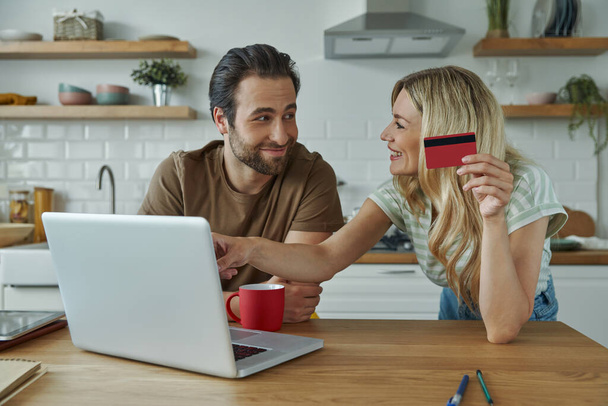Cheerful young couple using credit card while shopping online from domestic kitchen - Foto, Bild
