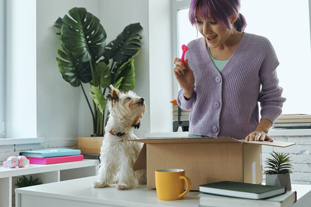 Beautiful young woman unpacking box while her cute dog sitting near her on the table - Фото, изображение