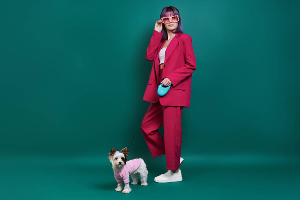 Glamourous young woman walking with little dog against green background - 写真・画像