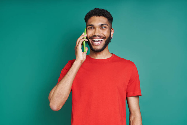 Ευτυχισμένος Αφρικανός μιλάει στο κινητό του και χαμογελάει ενώ στέκεται στο πράσινο φόντο - Φωτογραφία, εικόνα