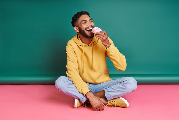 Cheerful African man enjoying doughnut while sitting against colorful background - Foto, imagen