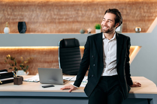 Позитивный профессиональный успешный кавказский исполнительный директор, бизнес-лидер, генеральный директор компании, в стильном деловом костюме, стоит у своего стола в современном офисе, смотрит в сторону, счастливо улыбаясь - Фото, изображение