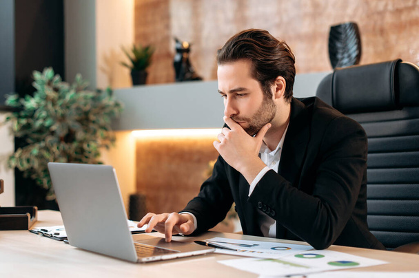 Сосредоточенный успешный кавказский умный бизнесмен, менеджер, бизнес-лидер, сидит на рабочем месте в офисе, внимательно смотрит на экран ноутбука, работает над бизнес-планом для нового проекта - Фото, изображение