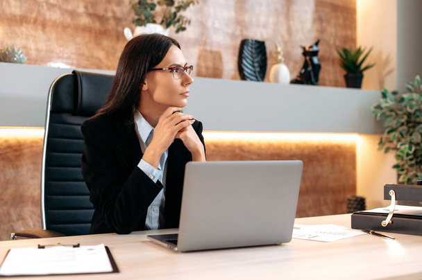 Pensive caucasian successful smart businesswoman with glasses and in a business suit, company financial manager, sits at a desk in the office, looks away, thinks about a project, plans a business plan - Photo, image