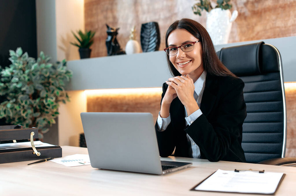 Портрет великолепной умной успешной кавказской брюнетки бизнес-женщины в очках, генерального директора компании, топ-менеджера, сидящего за столом в современном офисе, смотрящего в камеру, положительно улыбающегося - Фото, изображение
