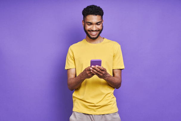 Handsome African man using smart phone and smiling while standing against purple background - Foto, imagen